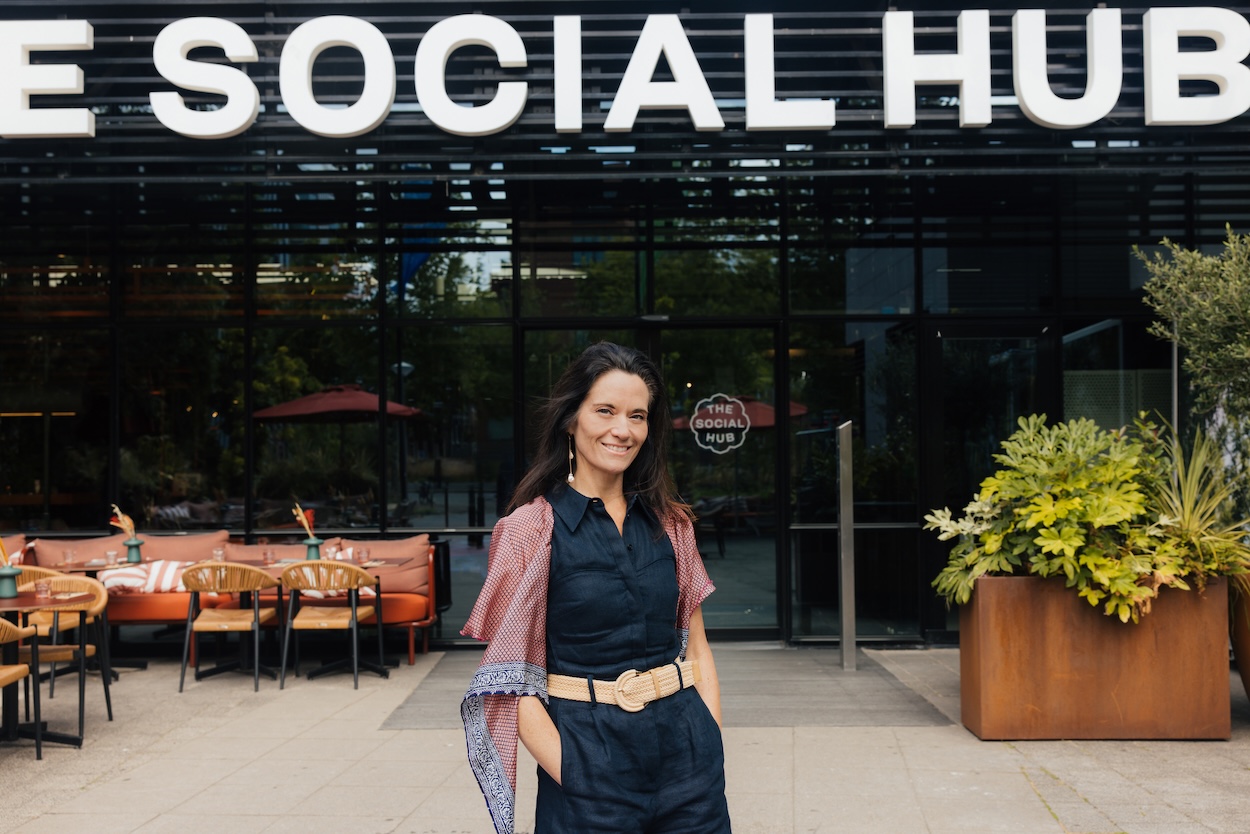 A smiling woman with her hands in her pocket in front of a building with a social hub sign