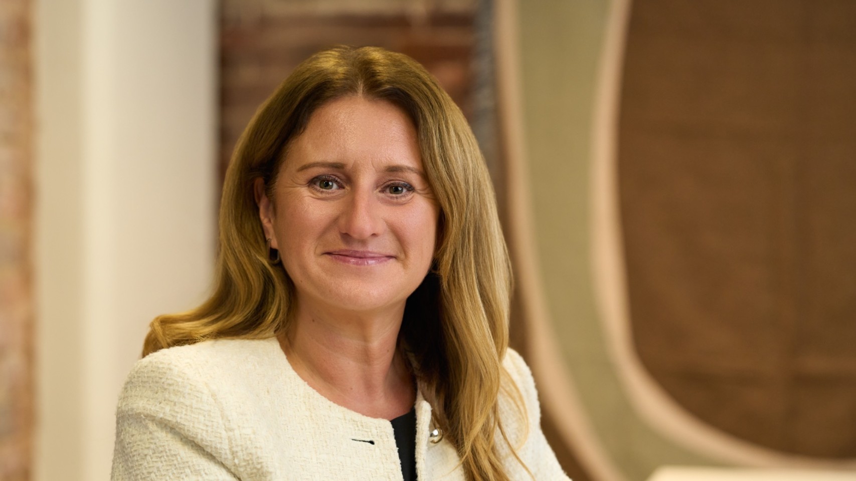 Charlotte Richmond sits at a table in a bright room, wearing a cream blazer and black top.