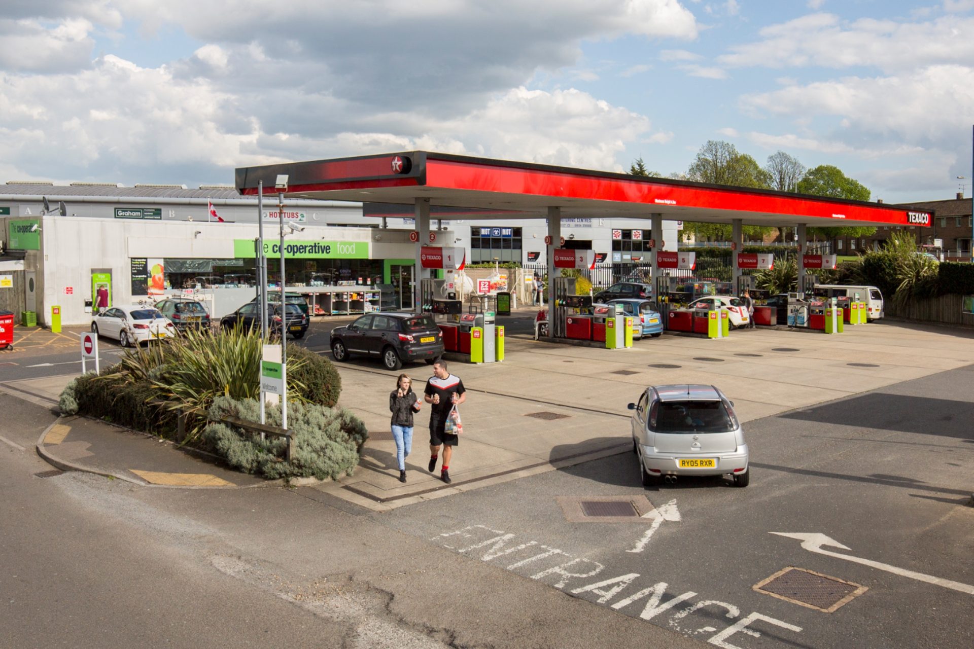 Rendered petrol station with a red canopy, cars on the forecourt and two people walking nearby.