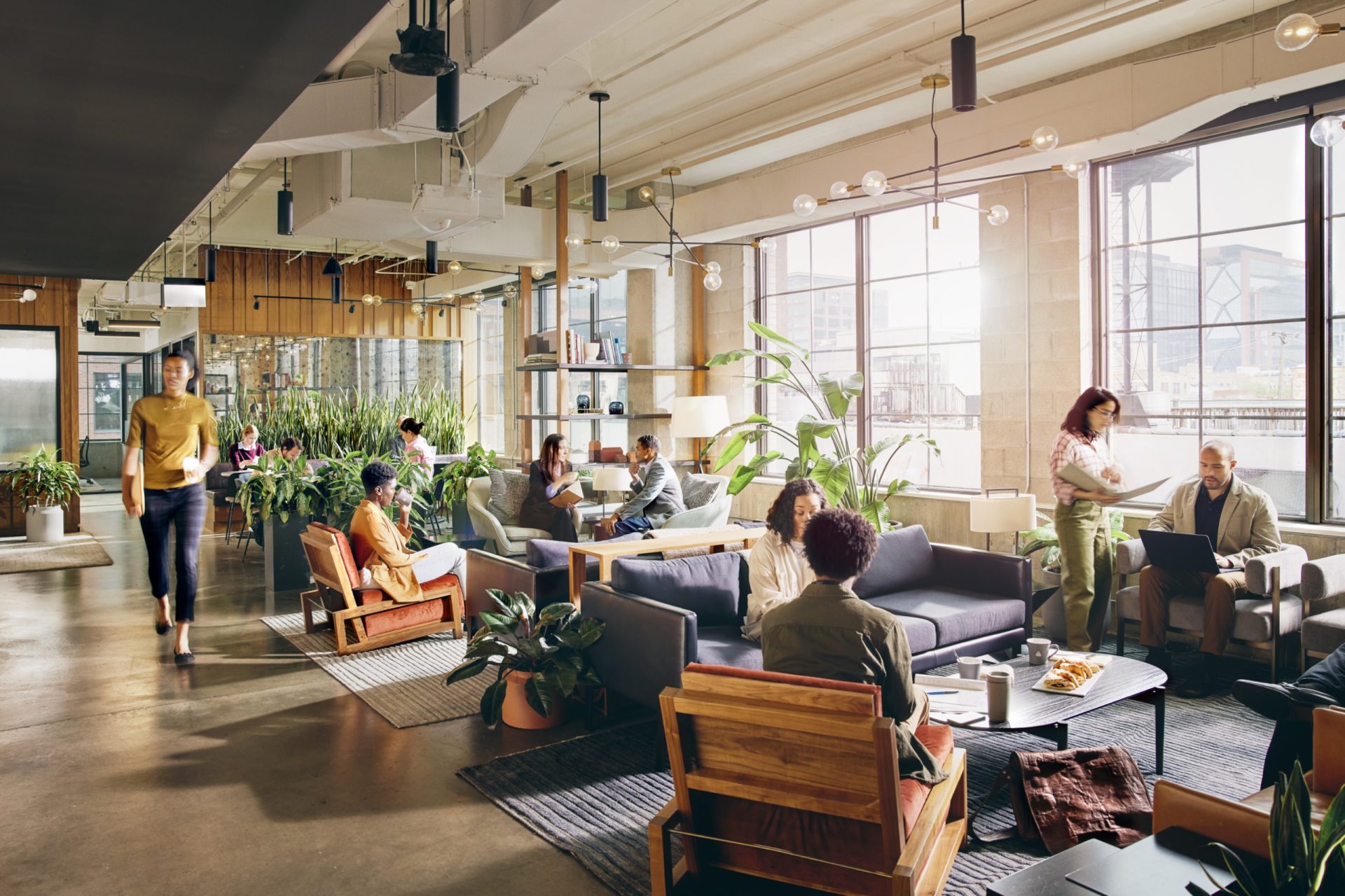 People work and talk in a bright office lounge with sofas, tables and large windows.