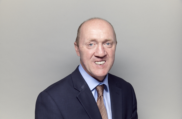 Smiling bald man in a dark suit and tie, pictured against a plain grey background