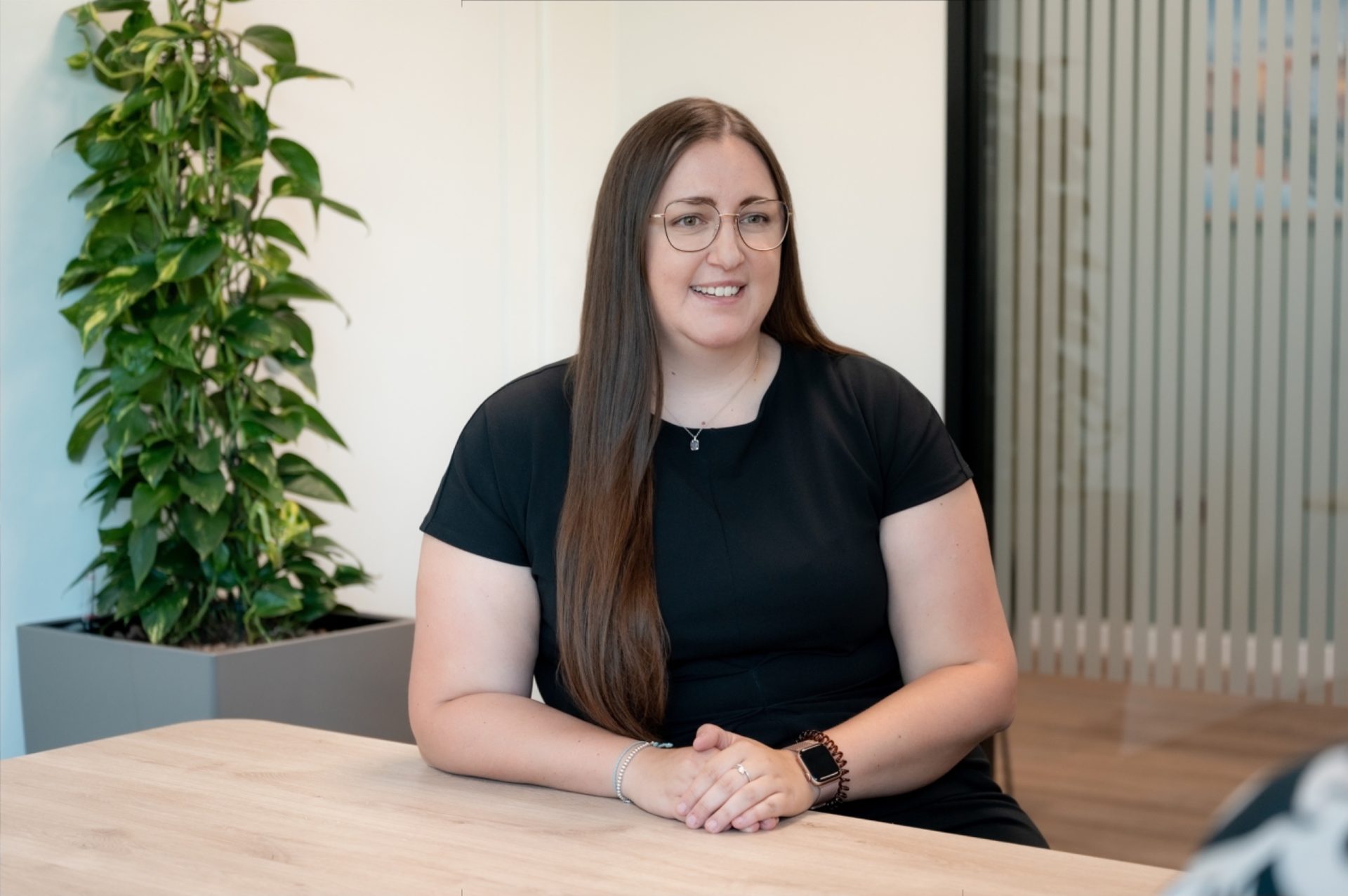 Woman in glasses sits at a table in a bright office with a potted plant nearby.