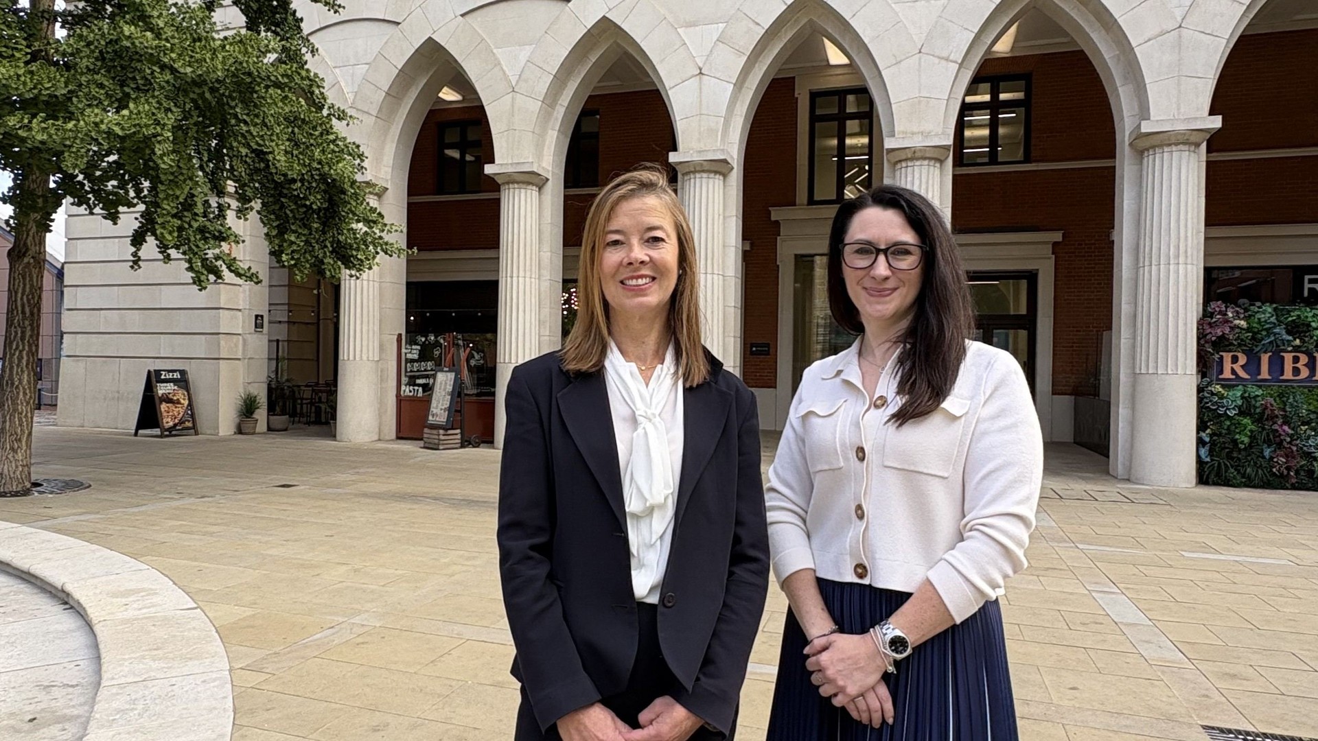 Emily Filer and Jenny Hemstock stand together outside a building with arched windows.
