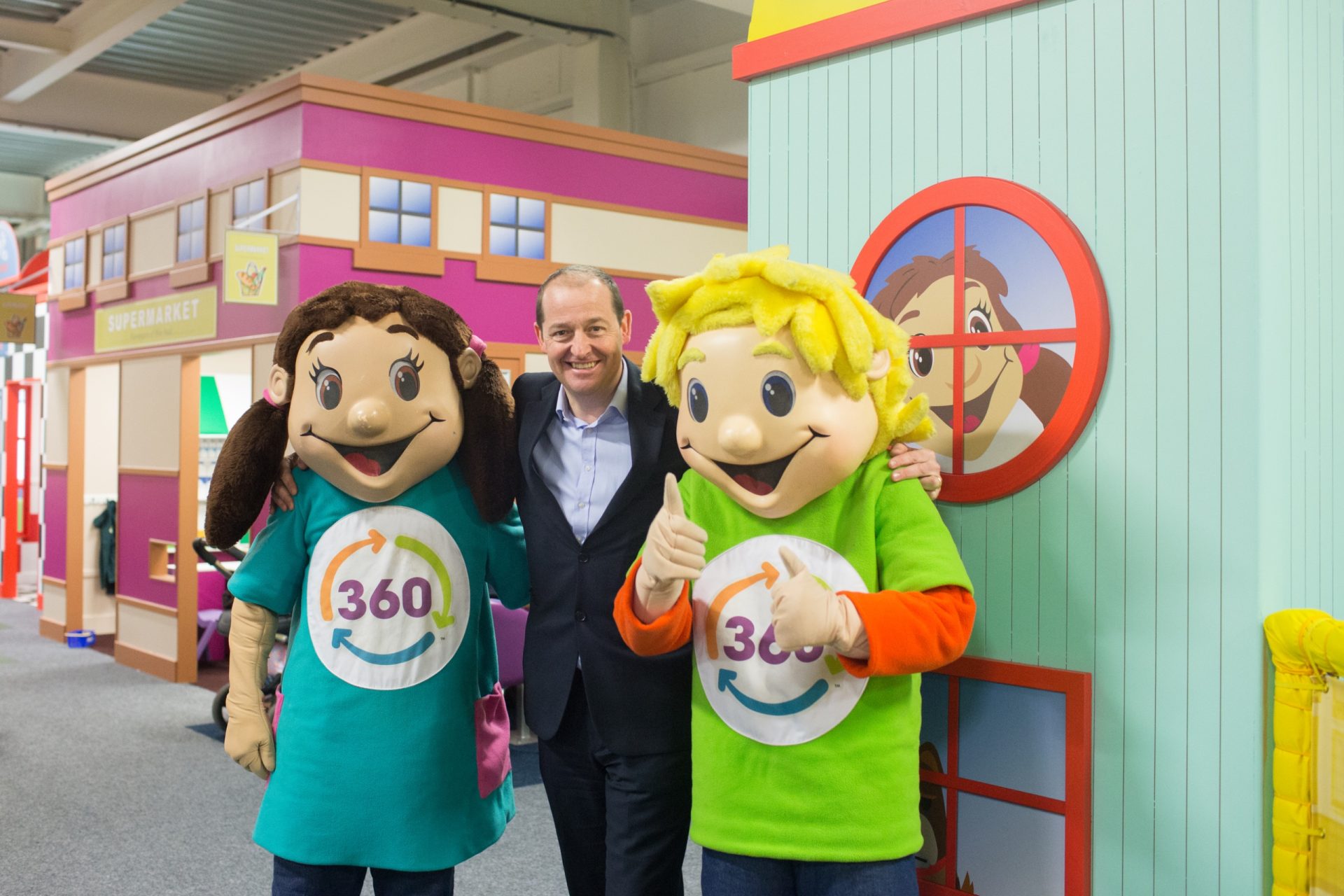 Two 360° mascots pose with a smiling man inside a brightly coloured children’s play area.