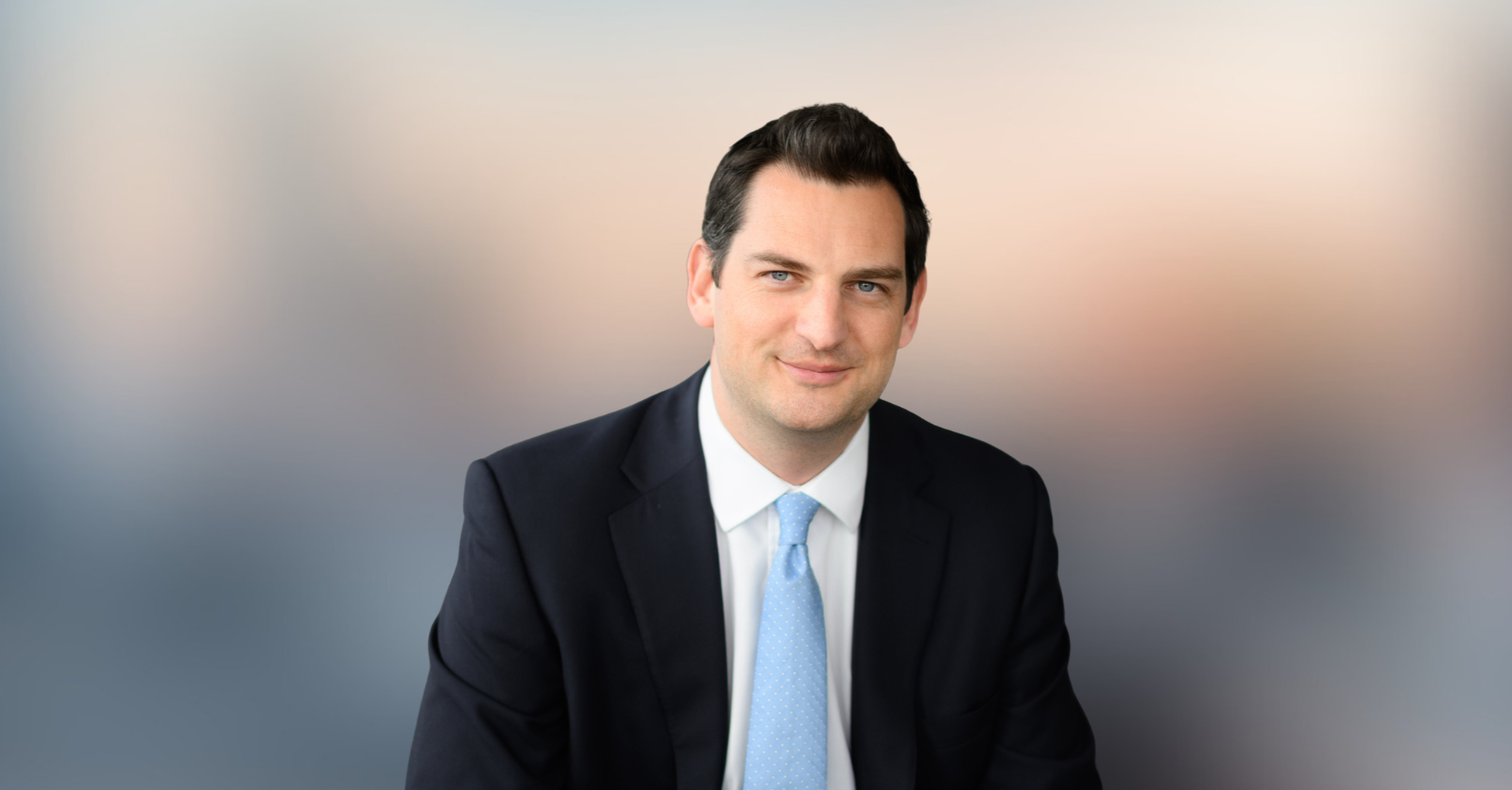 A dark-haired man in a navy suit and light blue tie poses against a softly blurred background.