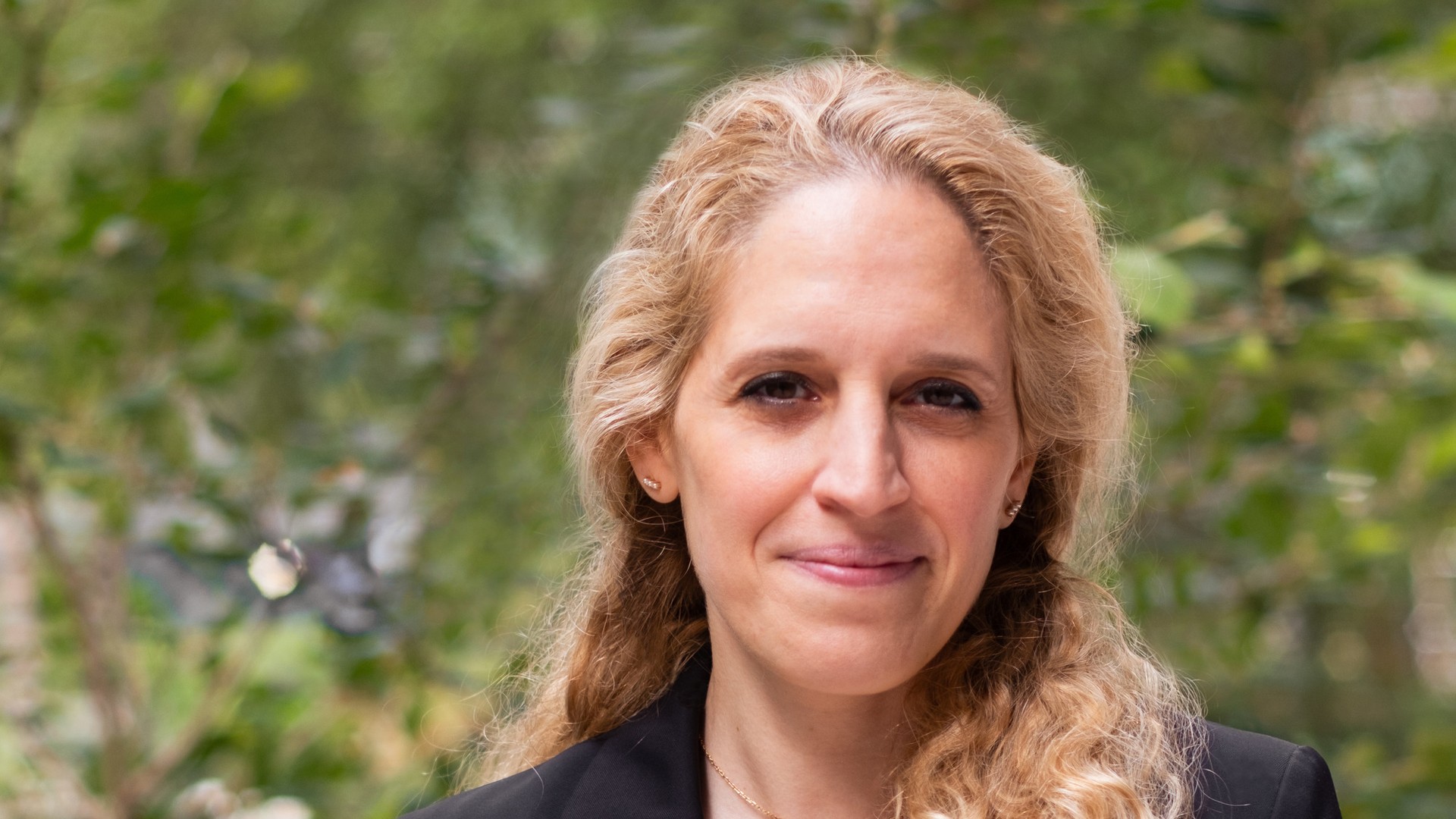 Mina Hasman OBE stands outdoors in a black blazer, with greenery behind her.