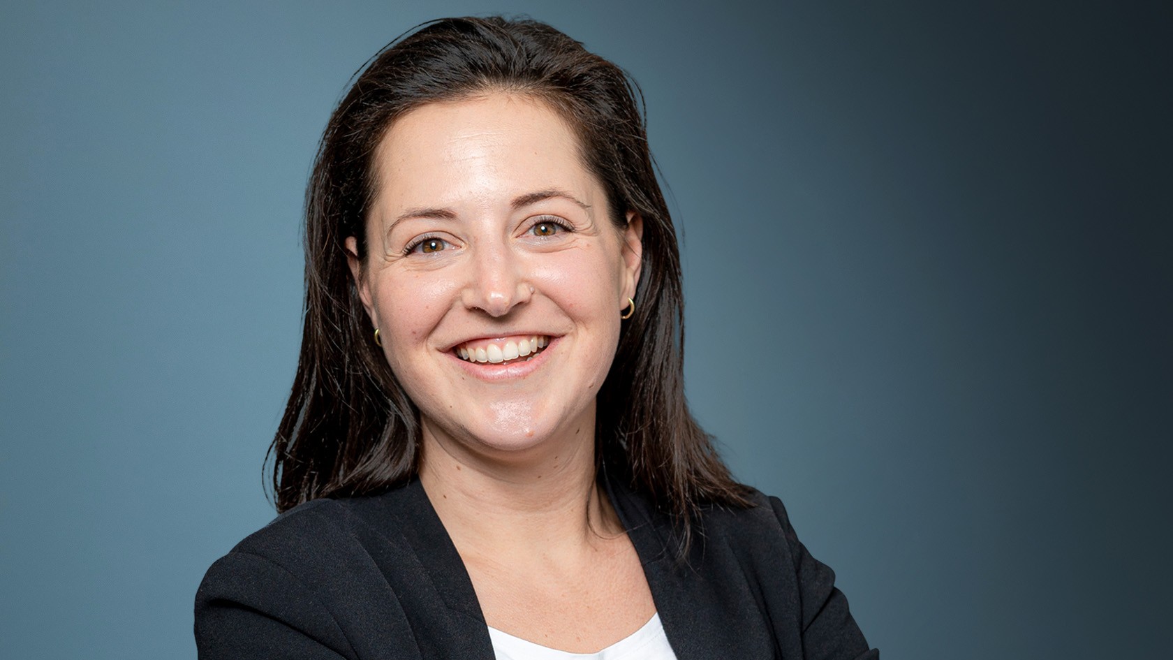 Smiling woman in a black blazer standing with her arms crossed against a blue background