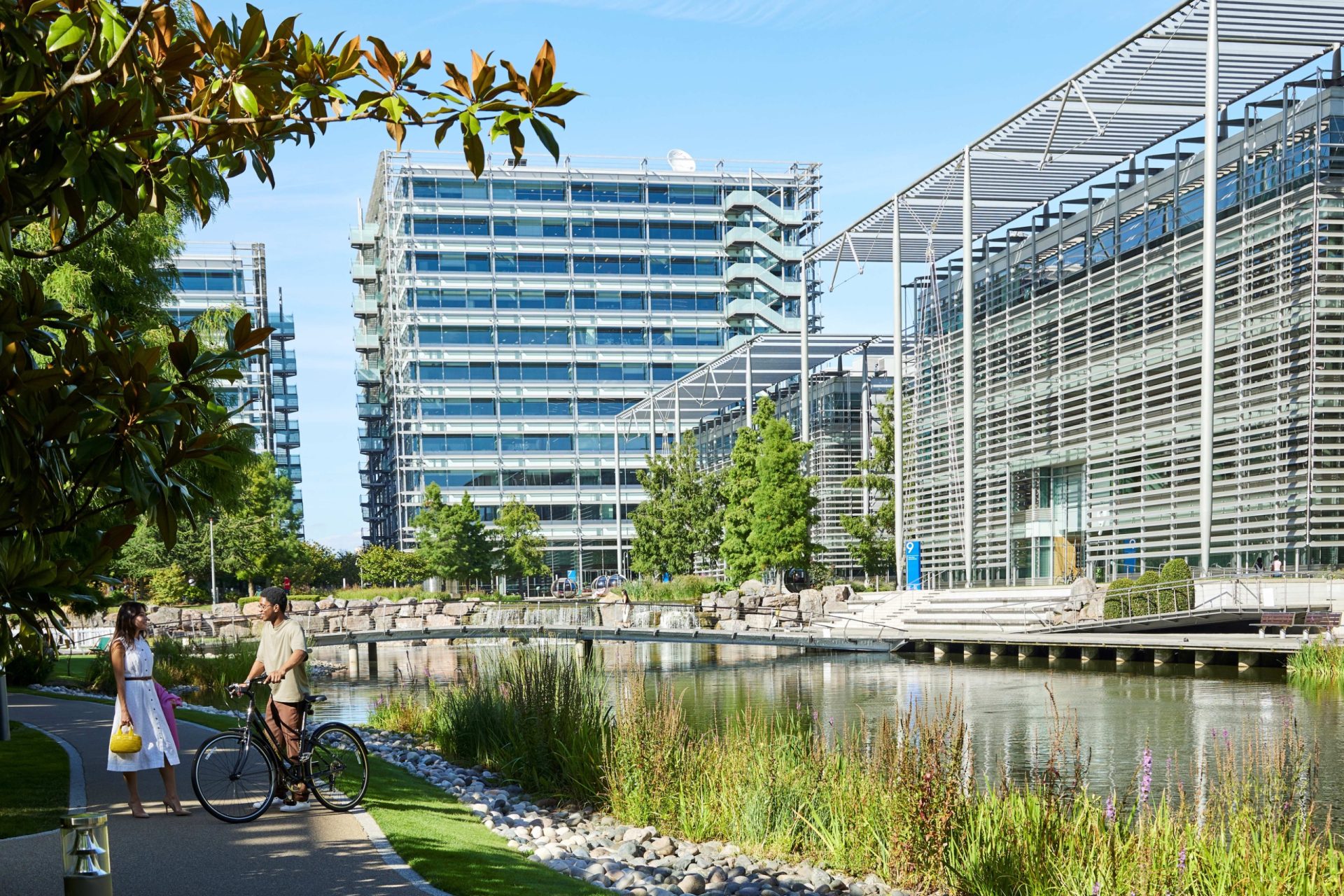 Modern glass office buildings at Chiswick Park beside a pond, with two people walking along the water.