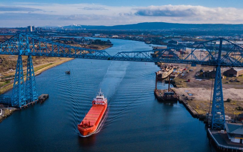 A red ship passing down a waterway under a large blue bridge