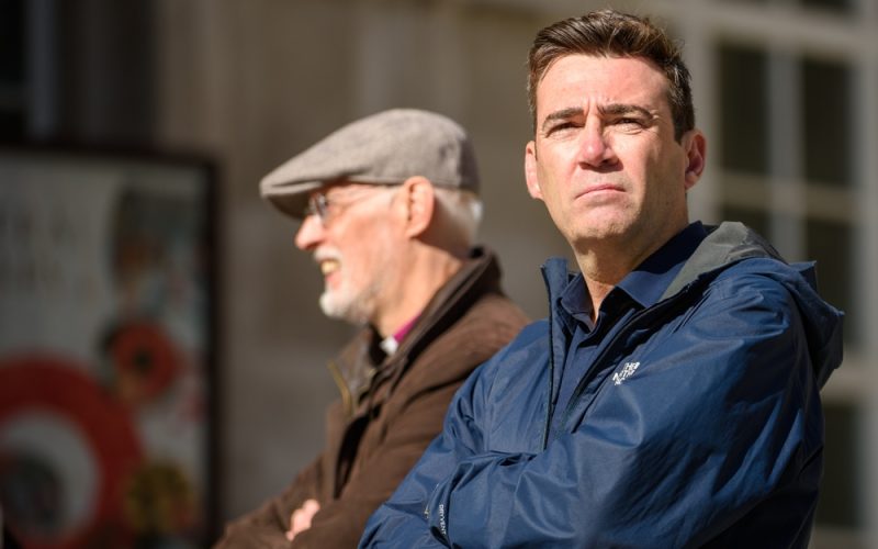 Andy Burnham wears a blue coat and looks off into the distance. He stands alongside an older man wearing a flat cap