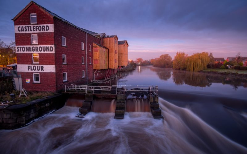 A fast flowing river with a brick built former mill building on the river banks