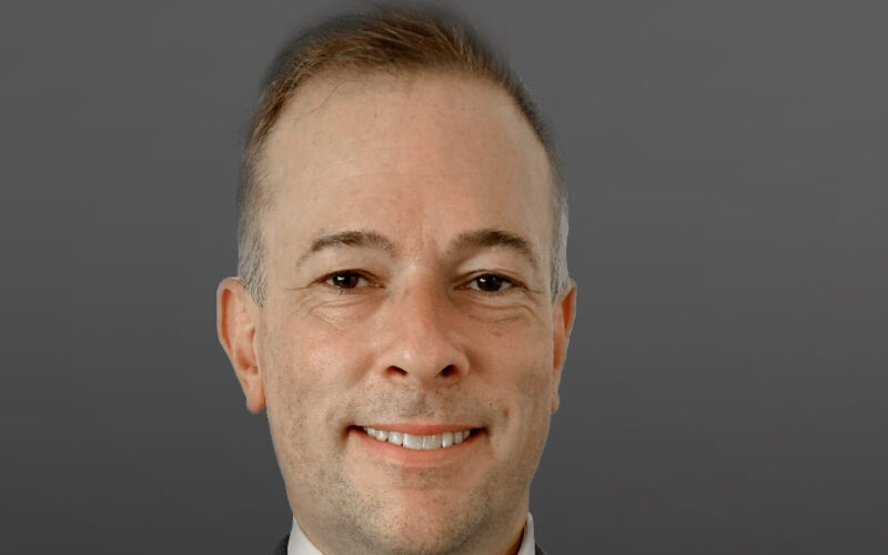 A smiling man with short hair wearing a dark suit jacket and a white shirt