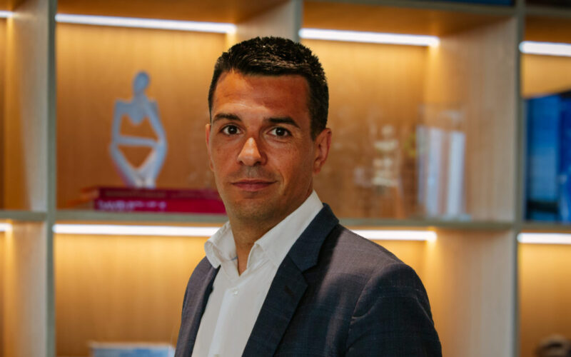 Picture of a man wearing a dark suit and white shirt with a glazed cabinet in the background