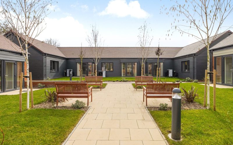 A connected series of grey wood panelled low rise buildings with a path and lawns in front