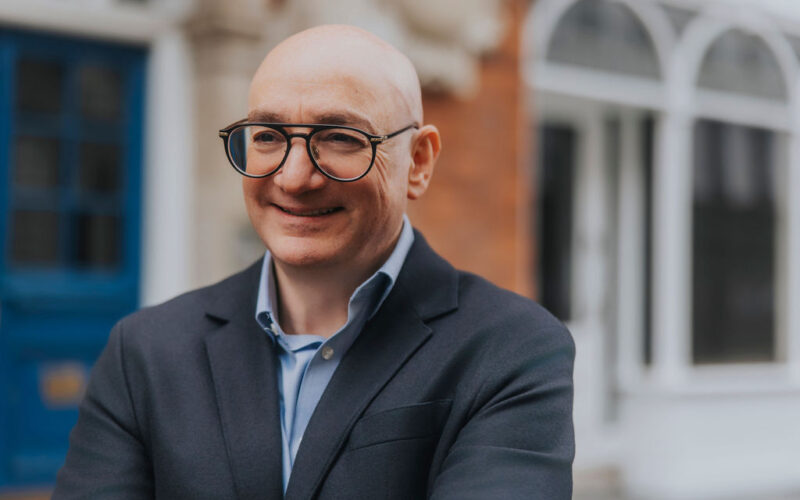 Ilan Goldman is a white, bald man wearing black rimmed glasses, a dark blue suit and a collared light blue shirt