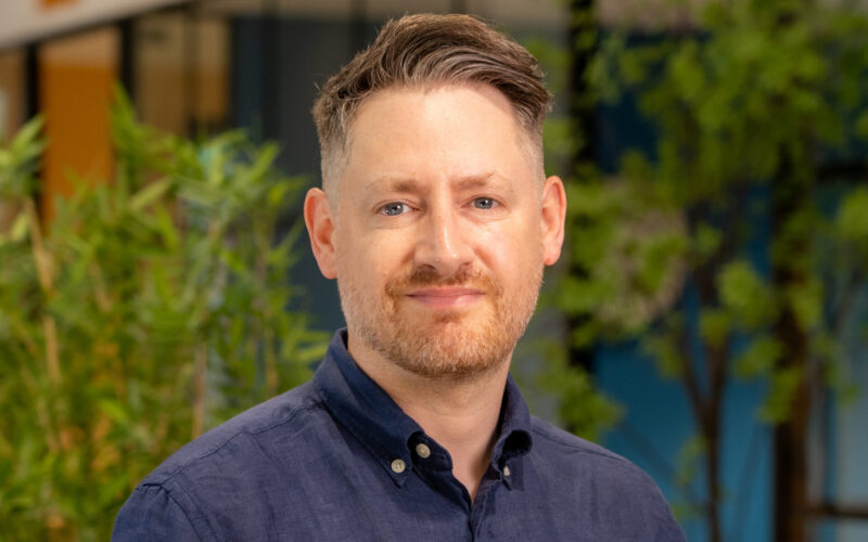 A while male wearing a dark blue collared shirt with trees in the background