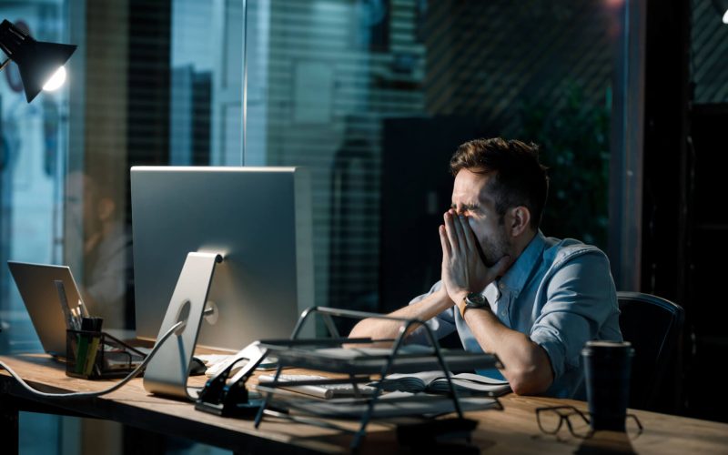 Man,Covering,Mouth,While,Yawning,At,Table,In,Dark,Office