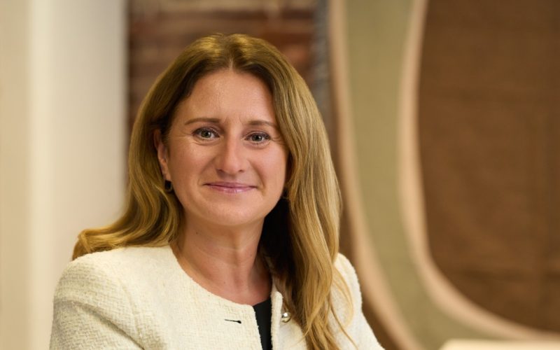 Charlotte Richmond sits at a table in a bright room, wearing a cream blazer and black top.