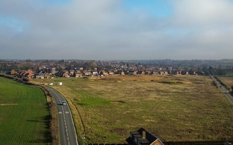 Aerial view of a rural road beside open development land and a suburban housing estate