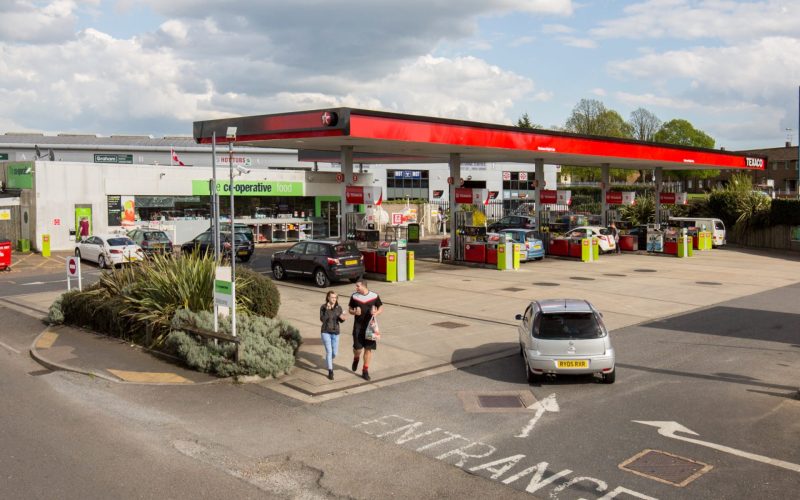 Rendered petrol station with a red canopy, cars on the forecourt and two people walking nearby.