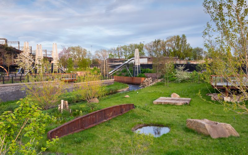 Grass-covered Mayfield Park site beneath railway arches, with a small water feature and surrounding buildings.