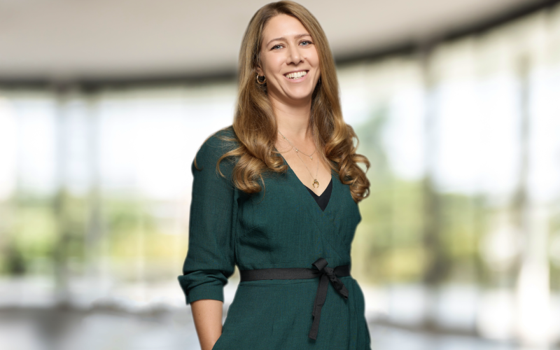 Alex Griffiths standing in a dark green dress against a blurred window background.