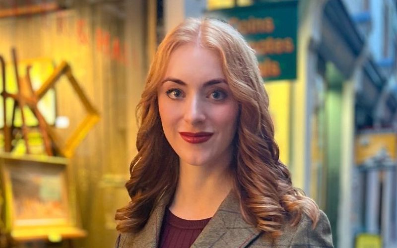 Saskia Jiggens stands in a grey checked blazer in a brightly lit indoor setting.