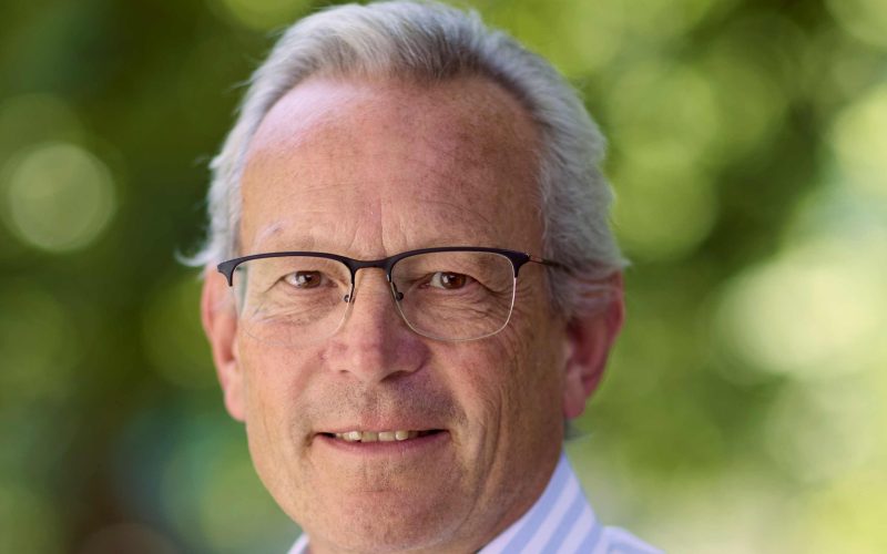 John Adams, chair of The Sirius Hotel Group, wearing glasses and a striped shirt against a green background.