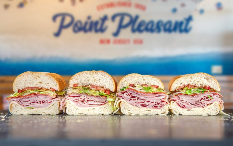 Four Jersey Mike’s-style subs filled with sliced meats, cheese, lettuce and onions, with a Point Pleasant sign behind them