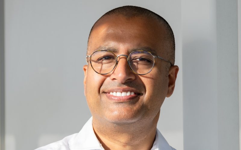 Kamran Tyler-Hussain, wearing glasses and a white shirt, smiles at the camera.