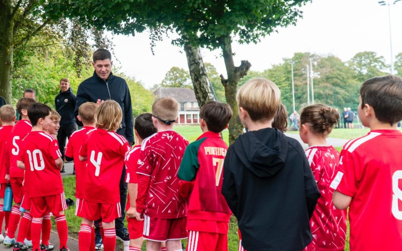Steven Gerrard stands with young footballers in red kits on a school sports field.