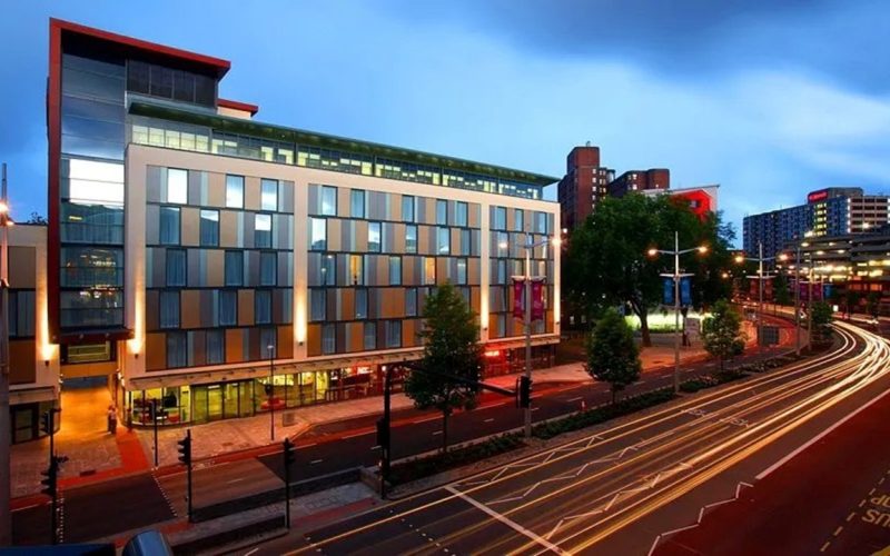 Modern hotel building beside a curving road at dusk, with city buildings and streetlights in the background.