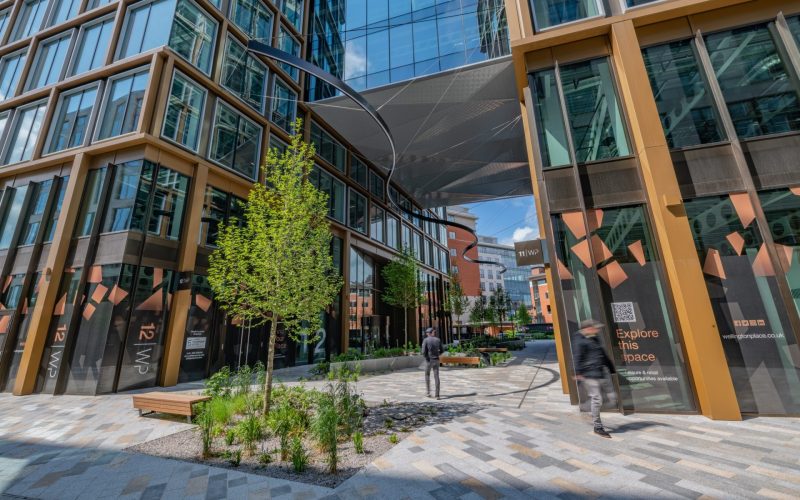 Modern commercial buildings lining a landscaped pedestrian plaza with people walking between offices