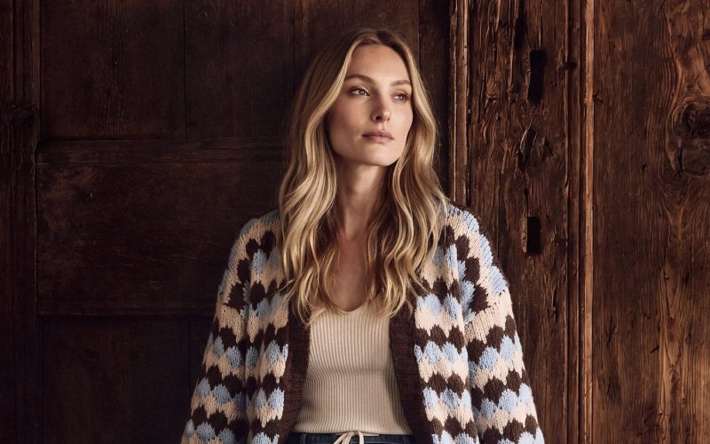 Woman in a patterned cardigan, white top, jeans and boots sitting on a rustic wooden bench