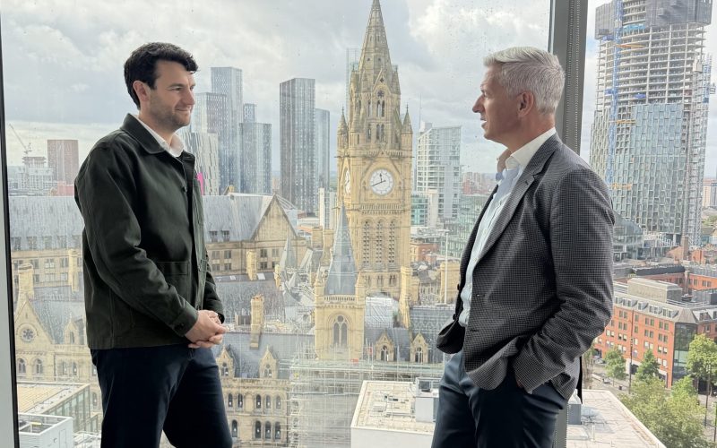Two men stand by a window overlooking Manchester, with the Town Hall clock tower behind them.