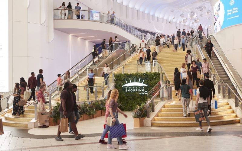 Shoppers walk through a multilevel indoor shopping centre, with the ONLY storefront visible beneath the escalators.