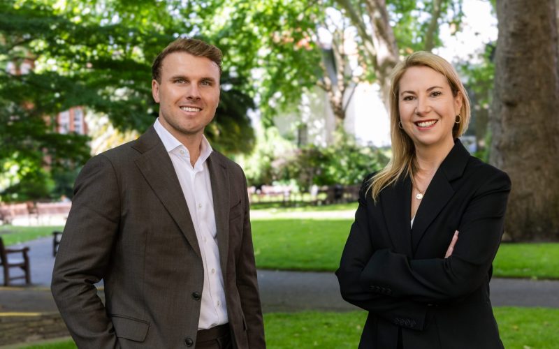 Henrik Orrbeck and Jessica Hardman stand outdoors in a leafy park, smiling at the camera.