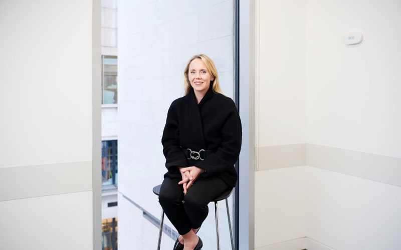 Gemma Kendall sits on a stool in a bright office, smiling at the camera.