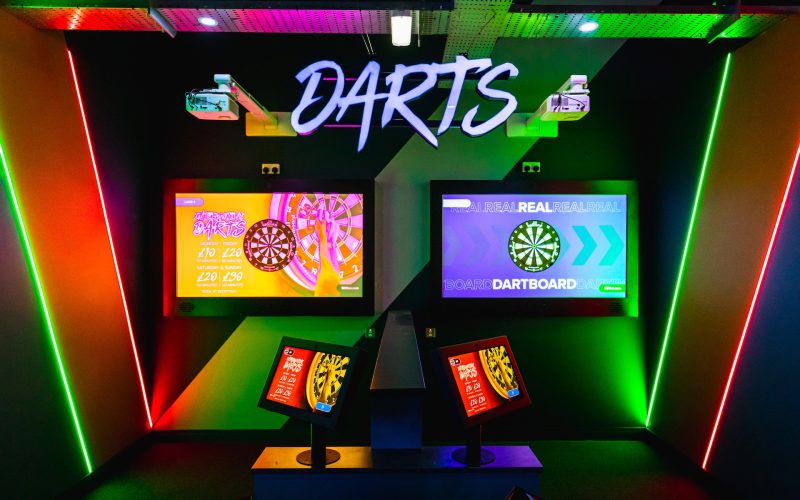Interactive darts setup with illuminated lanes, dartboards and scoring screens beneath a large “DARTS” sign