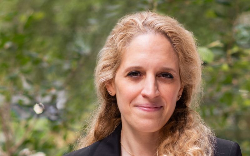 Mina Hasman OBE stands outdoors in a black blazer, with greenery behind her.