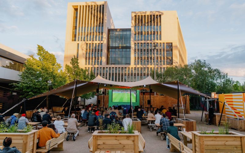 People gather at outdoor tables beneath a covered terrace, with Riverside East's modern building behind them.