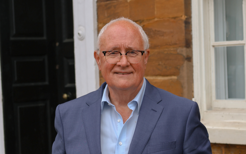 Sir Michael Lyons, wearing glasses and a blue suit jacket, seated in front of a wooden door and window.