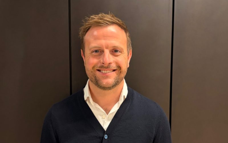 Jack Dukes stands smiling in a dark cardigan and white shirt against a paneled wall.