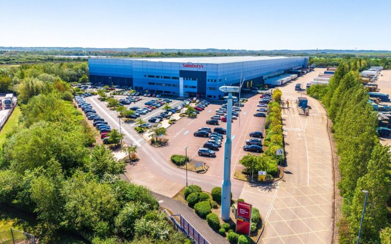 Aerial view of Sainsbury’s blue distribution centre in Bedford, with car parks, loading yard and surrounding trees.