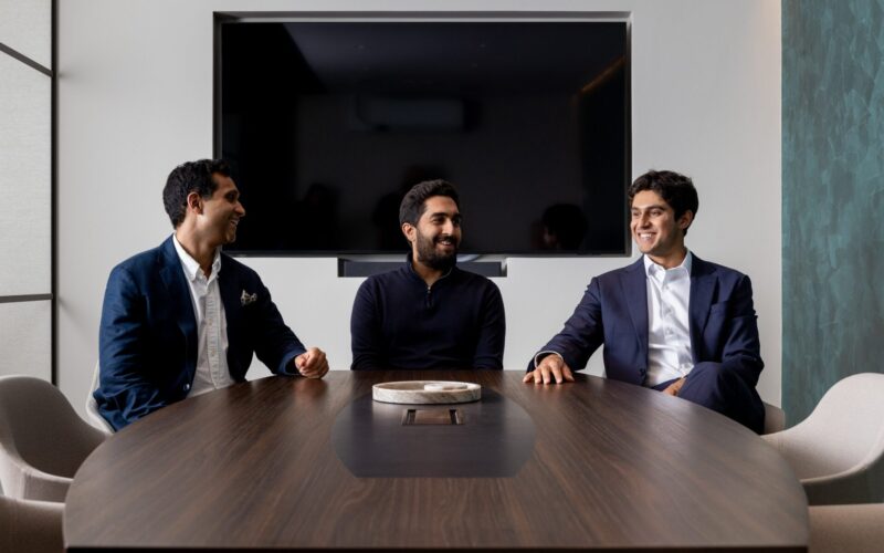 Three smartly dressed men sat around a table with a large TV screen behind them