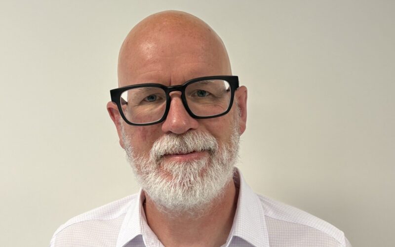 A bearded, bald white man wearing black rimmed glasses and a white shirt