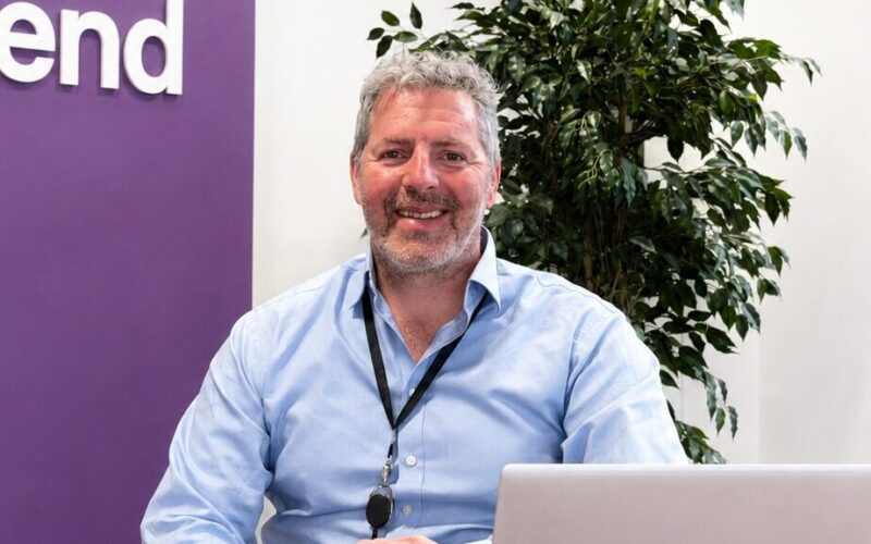 Picture of a smiling man wearing a blue shirt sat behind a table with a computer