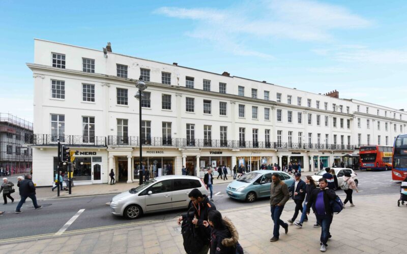 A parade of shops housed in a terraced building with a white facade with cars and pedestrians in the foreground