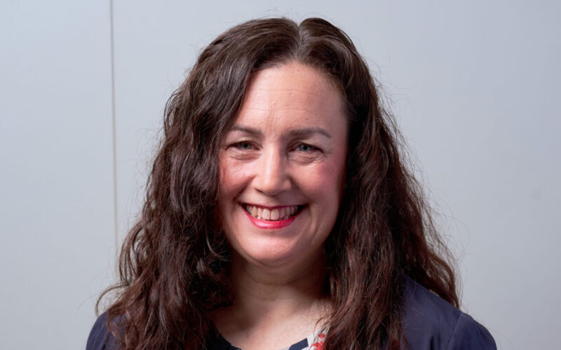 A smiling woman with dark long curly hair wearing a blue jacket and blue dress with a flower print