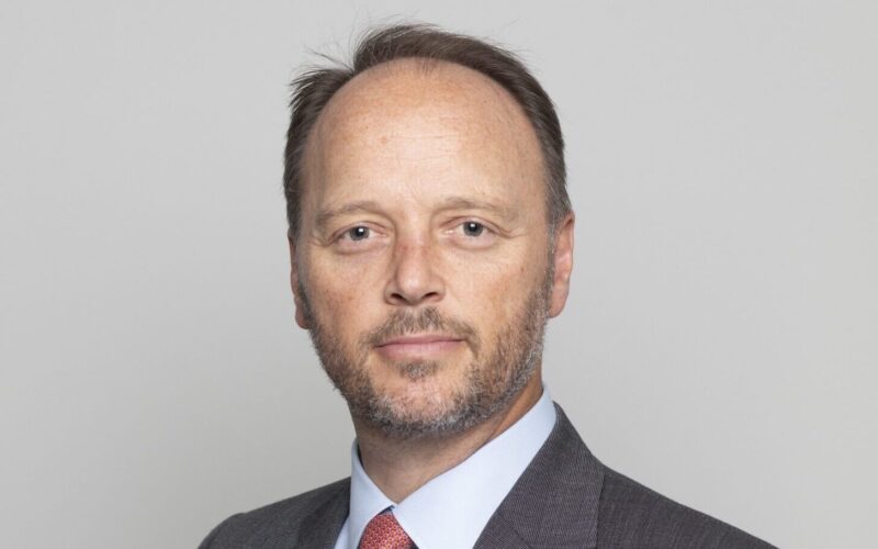 A white male wearing a grey suit jacket, a light shirt and a red tie